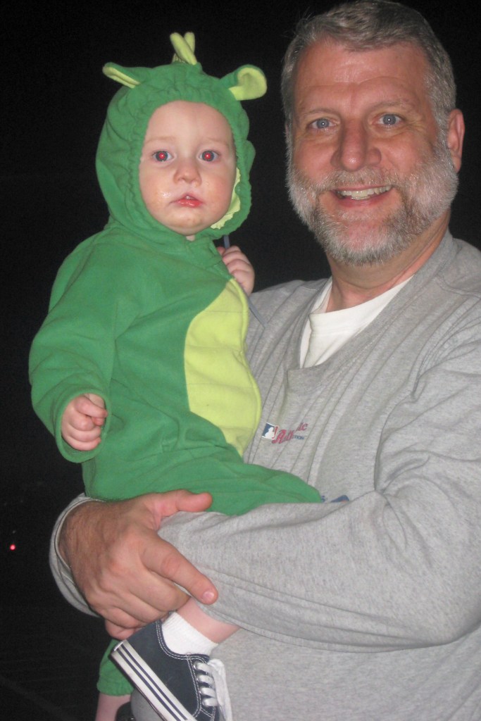 A smiling man holding a baby dressed in a green frog costume. The baby appears to be a toddler with a playful expression. The background is dark, suggesting an evening setting.