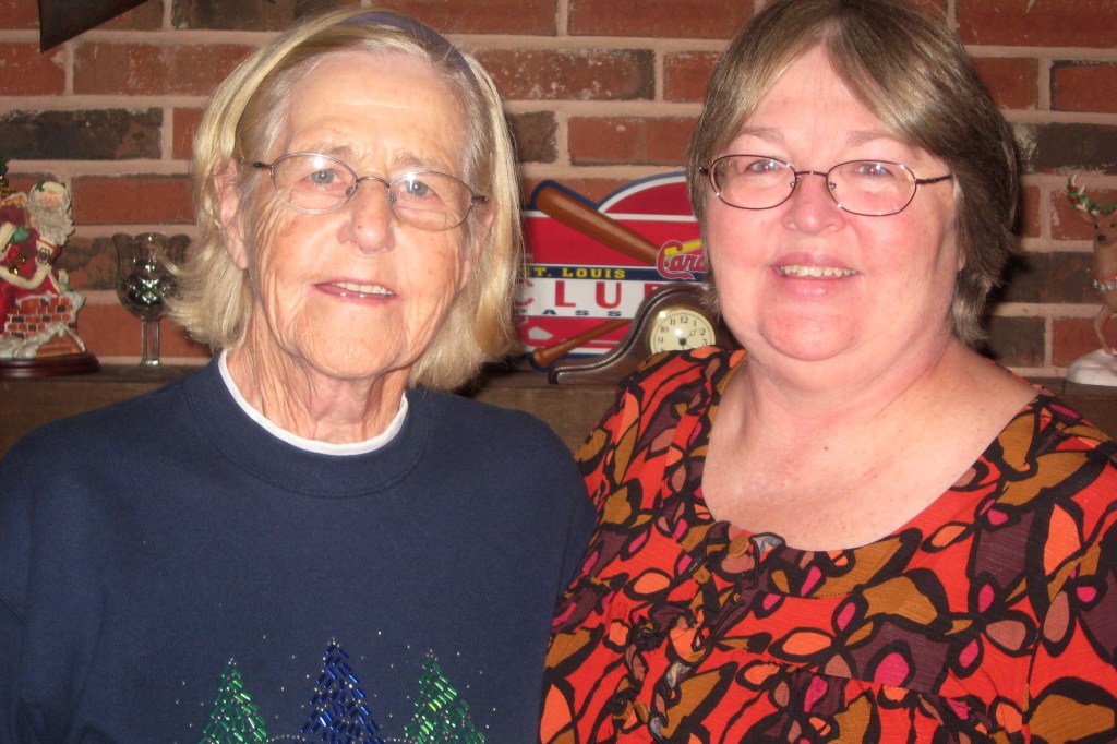 An elderly woman in a dark sweater with a decorative design, standing next to a middle-aged woman wearing a colorful patterned blouse, both smiling in front of a brick wall with various decorations.